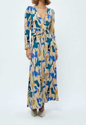 Femme portant une longue robe beige avec des motifs de feuilles bleues, vertes et jaunes, et des chaussures blanches à bouts pointus.