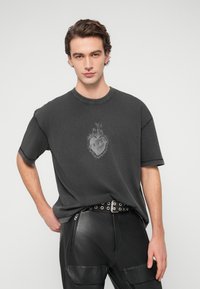 Σκούρο γκρι κοντομάνικο μπλουζάκι με γραφικό σχέδιο καρδιάς στο κέντρο και εμφανείς λεπτομέρειες ραφής. Συνδυασμένο με μαύρο δερμάτινο παντελόνι.