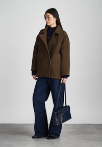Brown wool blend double-breasted coat with a textured finish, large black buttons, side pockets, paired with dark blue wide-leg jeans and a navy handbag.