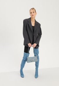 Junge Frau in übergroßem dunklem Blazer, schwarzem Outfit, blauen kniehohen Stiefeln, die eine kleine blaue Handtasche vor einem schlichten weißen Hintergrund hält.