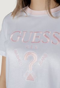 Balta medvilninė marškinių megztinys su rožine siuvinėta fraze "GUESS U.S.A." ir klaustuko dizainu. Yra rožinė megzta apykaklė.