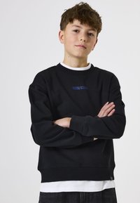 Sudadera negra con cuello redondo, con texto azul y mangas largas. Lleva puesta sobre una camisa blanca, con un ajuste relajado y puños acanalados.