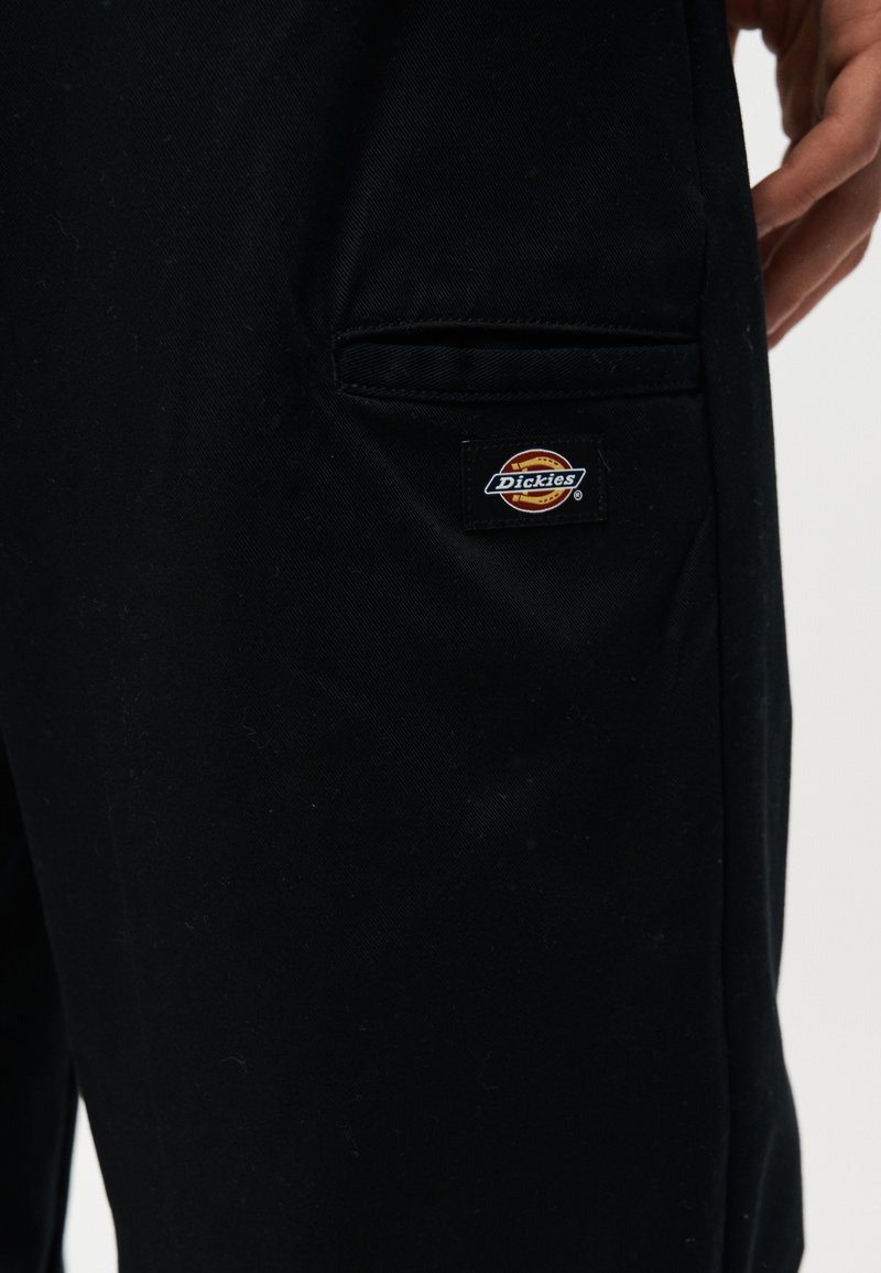Gros plan sur la poche d'un pantalon noir avec un patch logo Dickies sur le côté, une main est partiellement visible près de la poche.