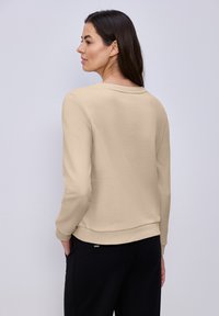 Femme aux longs cheveux foncés portant un sweat-shirt beige à manches longues et un pantalon noir, debout contre un fond gris clair, tournée vers la gauche.
