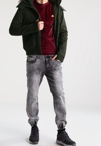 Mørkegrøn zip-up jakke, burgunder skjorte, grå tapered joggebukser og sorte sneakers med grå detaljer. Glatte teksturer og afslappet pasform.