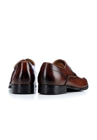 Zapatos de vestir de cuero marrón con una textura suave, puntera redondeada y suela de goma negra. La vista trasera muestra una costura mínima y un diseño clásico.