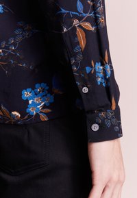 Mustariblommor med blå och bruna blommor på svart skjorta. Har en knappmanschett och en mjuk textur.
