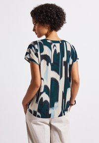 Model faces away, showcasing a patterned top in teal and cream hues, paired with light trousers. She stands against a neutral background.