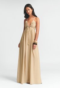 Długa, beżowa sukienka maxi z głębokim dekoltem w kształcie V, marszczonym biustem i regulowanym wiązaniem. Lekka tkanina swobodnie układa się do podłogi.