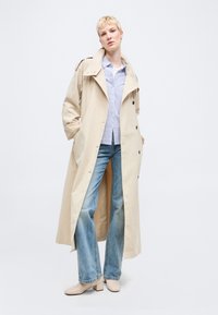 Άτομο που φοράει μακρύ μπεζ παλτό τρεντς πάνω από μπλε ριγέ πουκάμισο και φαρδιά μπλε τζιν, στέκεται με τα χέρια στις τσέπες του παλτού.