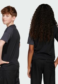 Schwarze sportliche T-Shirts mit kurzen Ärmeln, strukturiertem Stoff, mit einem schlichten Design und kontrastierenden Schultereinsätzen, getragen von zwei Kindern.