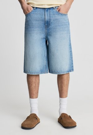 Shorts in denim azzurro chiaro con vestibilità ampia, vita media e orli risvoltati. Indossati con calzini bianchi a coste e scarpe slip-on marroni.