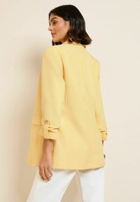 Kobieta z włosami do ramion w ciemnym kolorze, ubrana w żółtą marynarkę z podwiniętymi rękawami oraz białe spodnie, stojąca na beżowym tle.