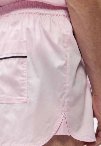 Ροζ ανοιχτόχρωμο σορτς με ελαστική μέση και τσέπη στο πλάι με μαύρο τελείωμα, φορεμένα από ένα άτομο με ορατό μπράτσο και χέρι.