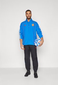 Modra trening jakna z rdečimi detajli in logotipom, črne hlače s stranskimi črtami, drži večbarvno nogometno žogo, športna prileganje.