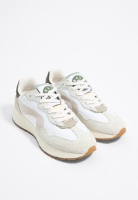 Sneaker bianche con accenti in suede beige, pannelli laterali in mesh e dettaglio del logo verde sulla linguetta. Lacci morbidi e suola in gomma marrone.