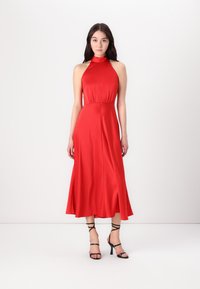 Robe dos nu rouge en tissu lisse, présentant un corsage ajusté et une jupe évasée. Portée avec des sandales à talons hauts noires à lanières.