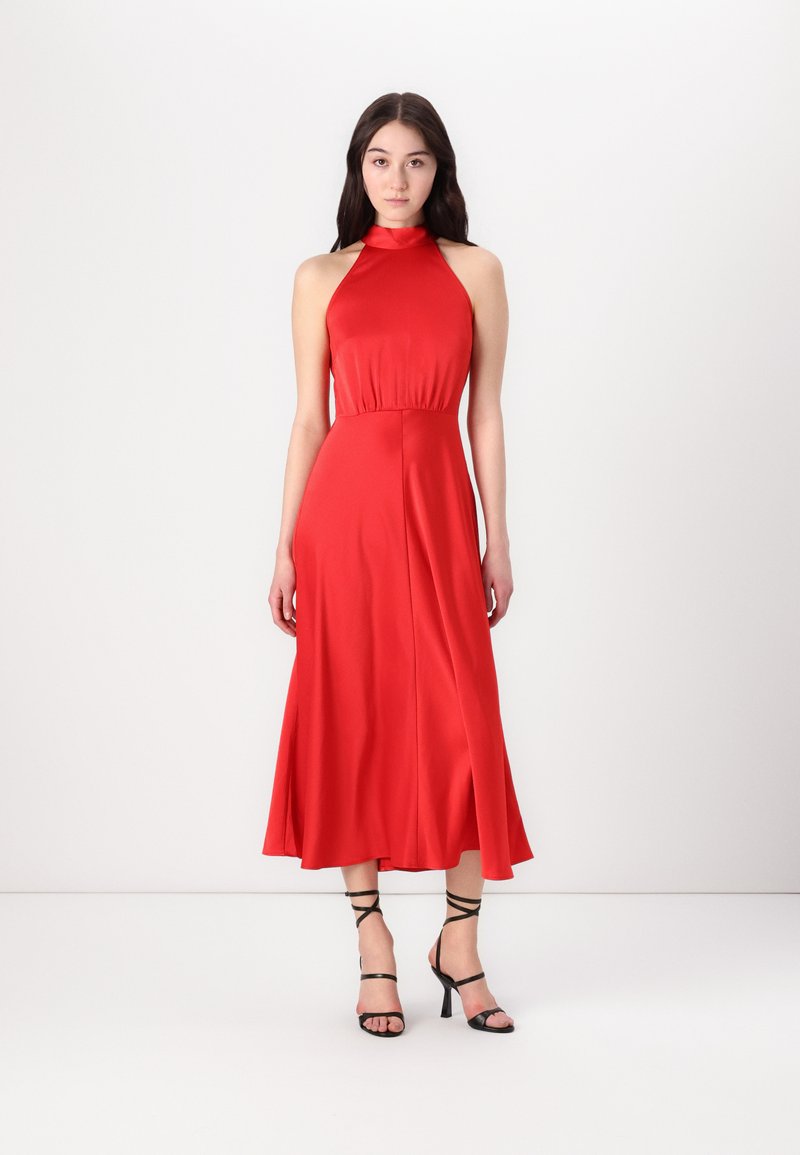 Robe dos nu rouge en tissu lisse, présentant un corsage ajusté et une jupe évasée. Portée avec des sandales à talons hauts noires à lanières.