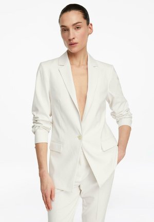 Kobieta ubrana w biały dopasowany żakiet z pojedynczym zapiętym guzikiem i pasujące białe spodnie, pozująca z jedną ręką w kieszeni.