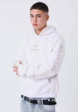 Joven con cabello corto y oscuro que lleva una sudadera blanca con rayas estampadas en las mangas y vaqueros azul claro con cinturón negro.