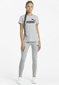 Szürke pamut póló fekete "PUMA" logóval, melyet passzoló, testhezálló szürke leggings-szel viselnek. A modellen fehér sportcipők vannak. Egyszerű, sportos dizájn.