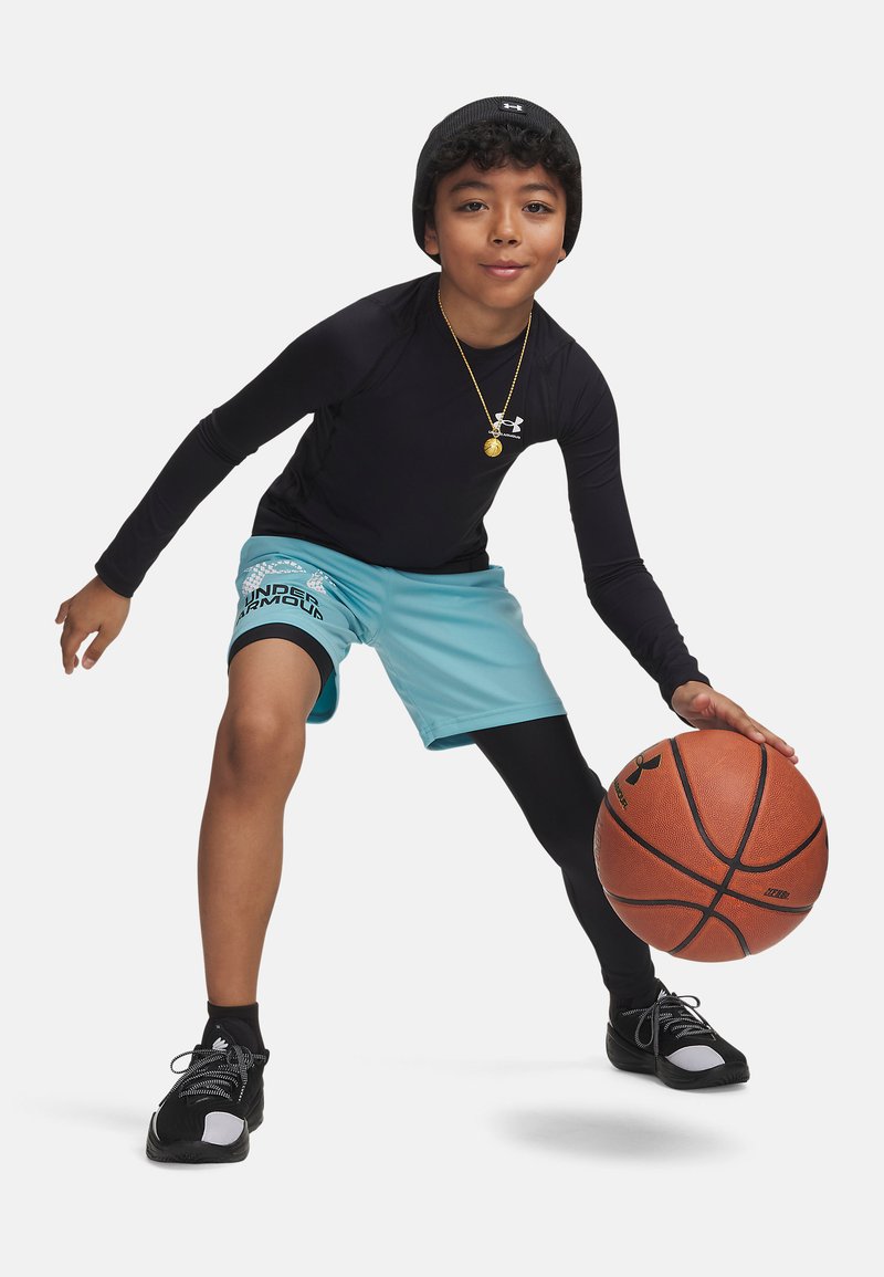 Jeune garçon portant un bonnet noir et une tenue de sport dribblant un ballon de basket orange avec un short bleu clair et des chaussures noires sur fond blanc.