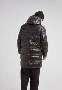 Abrigo acolchado negro con capucha, textura brillante, costuras horizontales y mangas largas, que llega hasta la mitad del muslo. Combinado con pantalones negros.