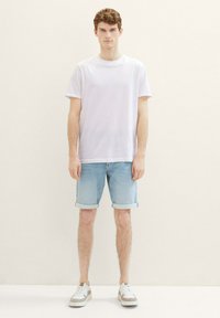 Camiseta blanca de algodón, pantalones cortos de mezclilla azul claro doblados en el dobladillo, y zapatillas gris-marrón con acentos blancos. Fondo liso, modelo de pie.