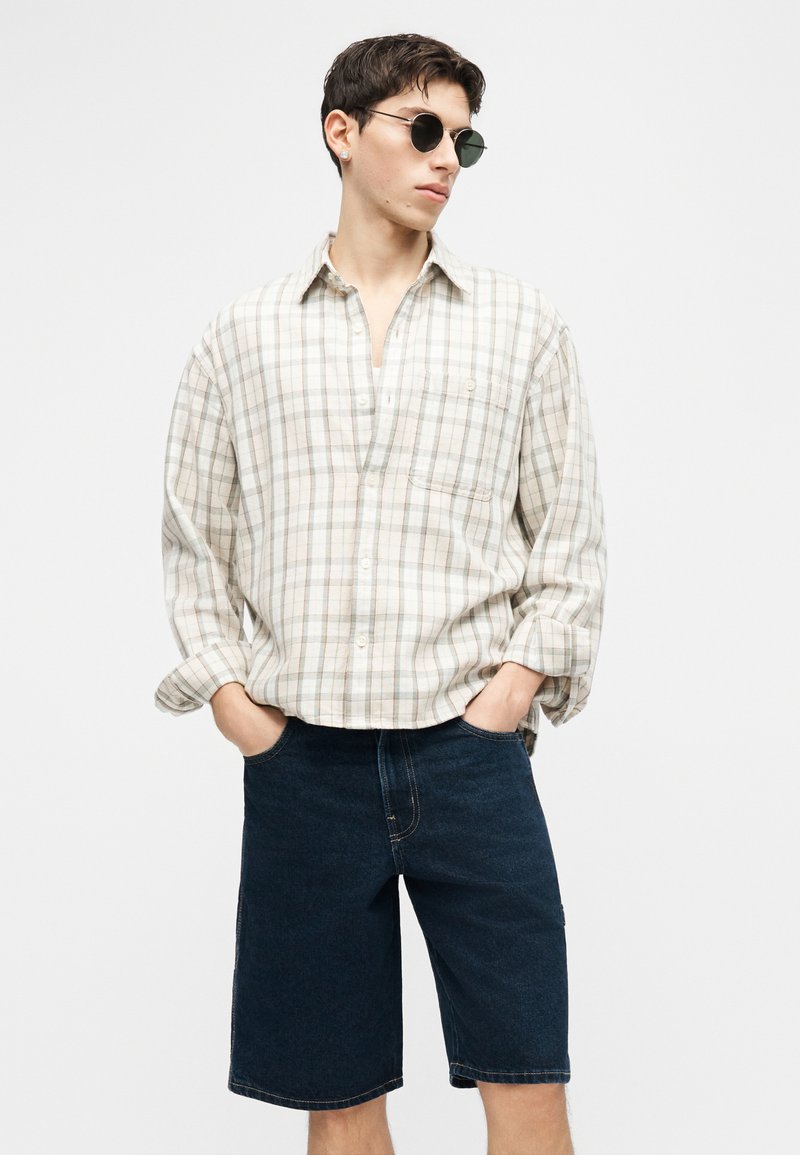 Giovane uomo con occhiali da sole rotondi, camicia beige a quadri con bottoni e pantaloncini di jeans scuri fino al ginocchio, mani in tasca, guarda di lato.