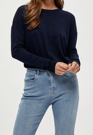 Femme portant un pull bleu marine à manches longues et un jean taille haute bleu clair, les mains doucement jointes devant elle.