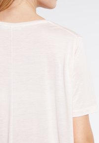 Kvinna som bär en enkel vit kortärmad skjorta, sedd bakifrån, med synlig axel- och nackprofil.