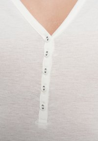 Λευκό, βαμβακερό πουκάμισο με ρίγες, διαθέτει ντεκολτέ σε σχήμα V και έξι ασημένια κουμπιά κατά μήκος του μπροστινού μέρους, με απαλό, υφασμάτινο φινίρισμα.