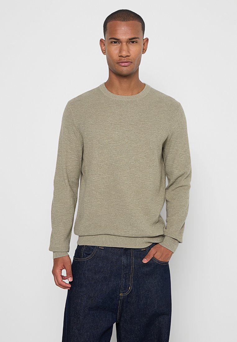 Licht olijfgroene gebreide trui met een ronde hals en geribde mouwen, gedragen over donkere spijkerbroeken. De textuur lijkt zacht en lichtjes gestructureerd.