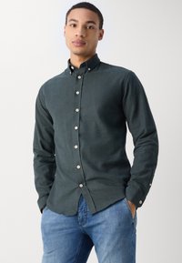Dark green button-up shirt made of textured fabric, featuring a collar and silver buttons, paired with blue denim jeans.
