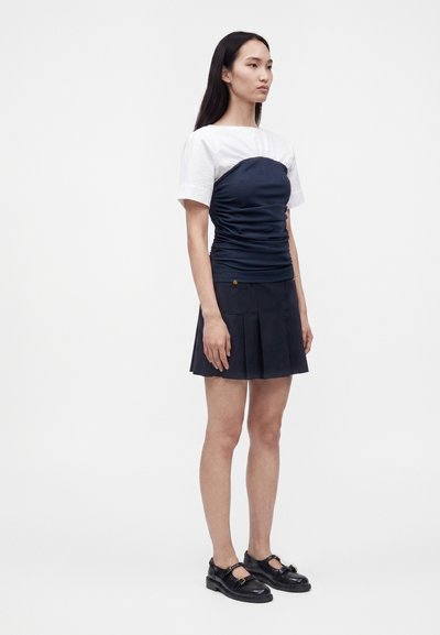 Jeune femme debout portant un haut blanc à manches courtes sous un bustier froncé bleu marine avec une jupe plissée bleu marine et des chaussures noires.