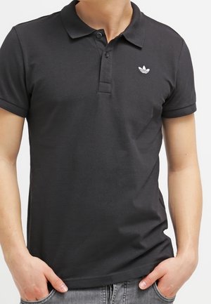 Homme portant un polo noir à manches courtes avec un petit logo Adidas blanc sur la poitrine gauche et les mains dans les poches d’un jean gris.
