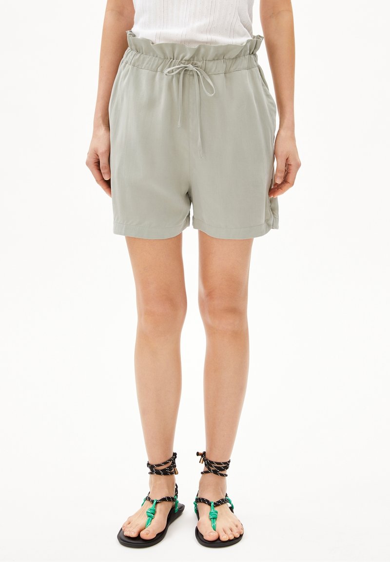 Personne portant un short vert clair à cordon de serrage et des sandales noires avec des brides vertes et noires aux chevilles, debout sur fond blanc.