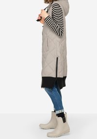 Puchowa kamizelka w jasnoszarym kolorze z kapturem, z ozdobnym pikowaniem w romby, bocznymi zamkami błyskawicznymi i czarnym prążkowanym wykończeniem. Nosi ją osoba na czarno-białym pasiastym topie.