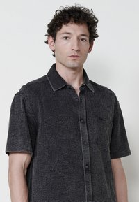 Camisa de cuello con mangas cortas en un gris oscuro, con un tejido texturizado, que presenta un cierre de botones y un bolsillo en el pecho.