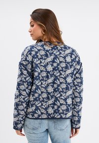 Veste matelassée marine avec motif floral en blanc et bleu clair, dotée d'un col rond et de manches à poignets. La vue arrière montre la texture et le design.