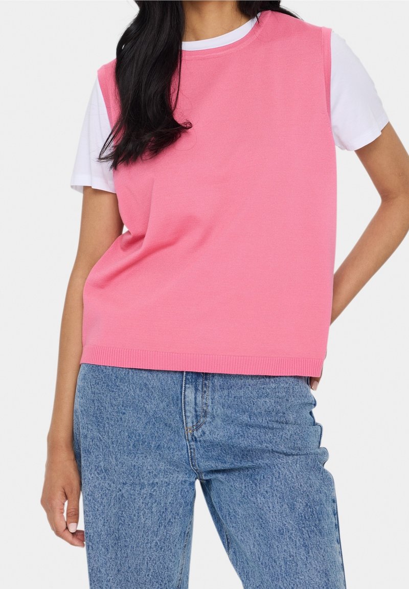 Kvinna som bär en ärmlös rosa sweatshirt över en vit kortärmad t-shirt och blå högmidjade denimjeans, står med ena handen på höften.
