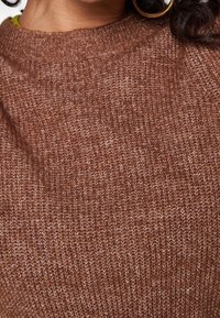 Gros plan d'une personne portant un pull en maille marron avec un col rond côtelé et une boucle d'oreille en or visible du côté droit.