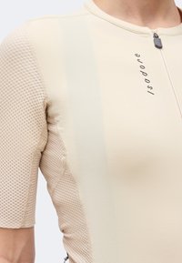 Κοντινή λήψη μπεζ ποδηλατικής φανέλας με υφασμάτινα μανίκια, μπροστινό φερμουάρ και λογότυπο "Isadore" στο στήθος.