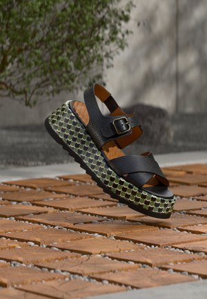 Črna usnjena sandal z križno prepletenimi trakovi, platformo z zeleno-belim vzorcem in rjavim oblazinjenim vložkom. Teksturiran podplat za oprijem.