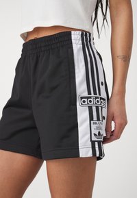 Bermudas deportivas negras con franjas laterales blancas y botones a presión. Presenta el parche del logo de Adidas y una tela texturizada con una cinturilla elástica.