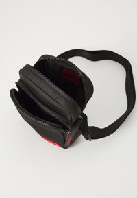 Black fabric crossbody bag with two zippered compartments, featuring a red logo tag and a textured exterior. Adjustable strap included.