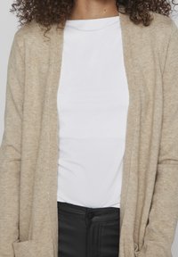 Beige kofta med öppen front, mjuk textur och långa ärmar, över en vit t-shirt, kombinerad med svarta byxor.