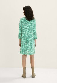 Grünes Blumenkleid mit Dreiviertelärmeln, knielangem Saum und lockerer Passform. Getragen mit braunen Stiefeln bis zum Knie.