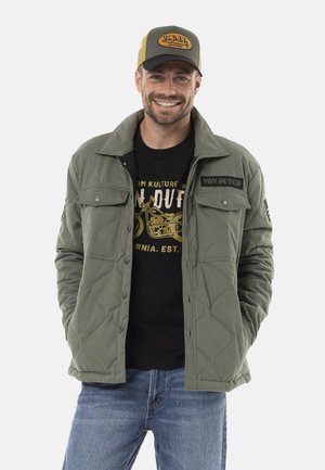 Homme souriant portant une veste matelassée vert olive, un t-shirt graphique noir avec une impression de moto, un jean bleu et une casquette beige Von Dutch.