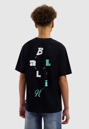 Tiener met krullend haar die een zwart T-shirt draagt met verspreide witte en groene letters die "Ballin" spellen op de rug, gecombineerd met een blauwe jeans.